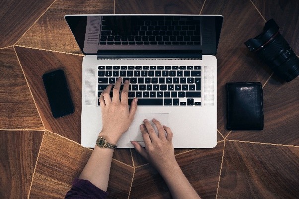 Overhead shot of a table with a Apple laptop open and two hands typing on it. There is a phone on the left side and a camera and wallet on the right side.