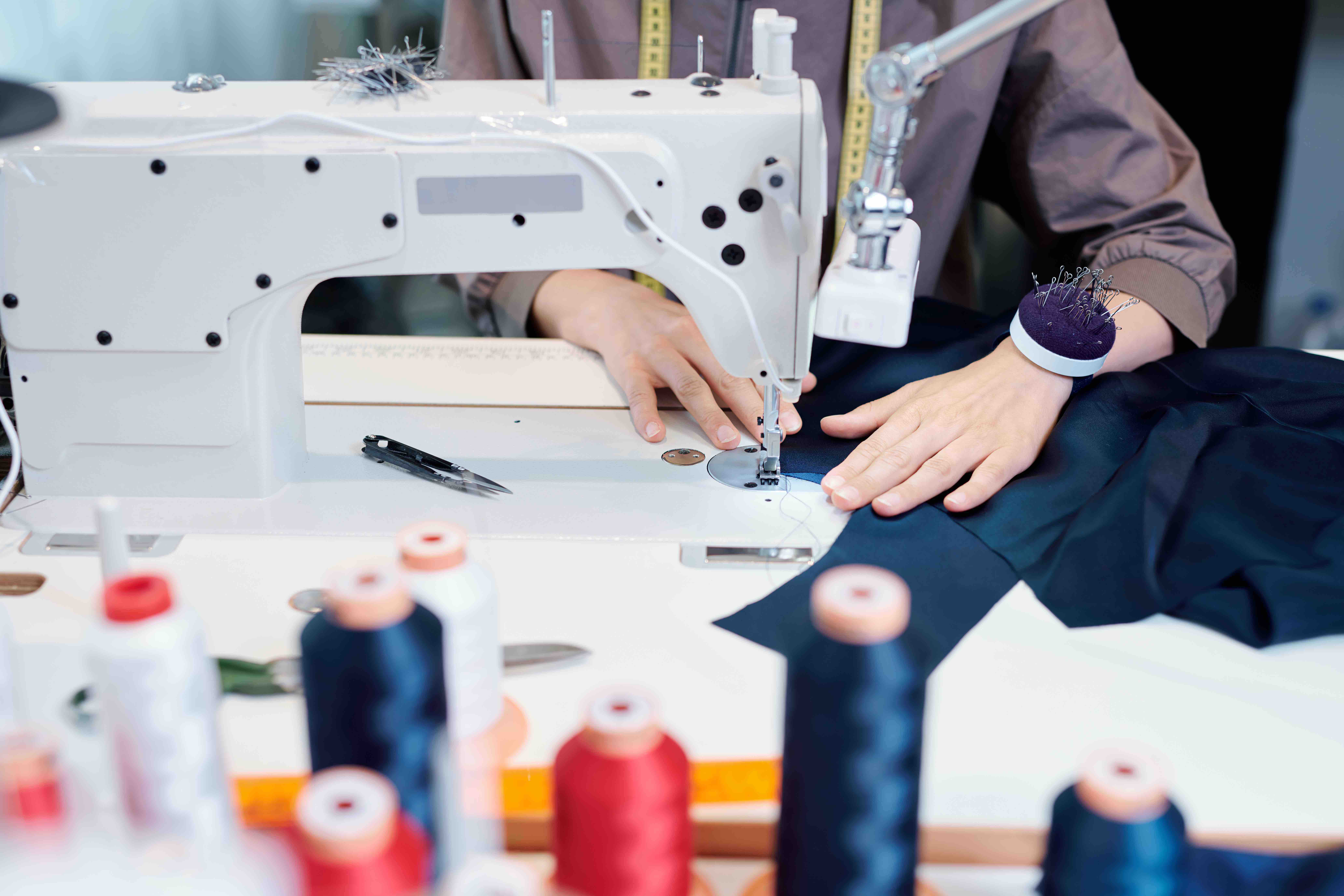 Female tailor hands moving piece of blue textile while sewing clothes by machine in workshop