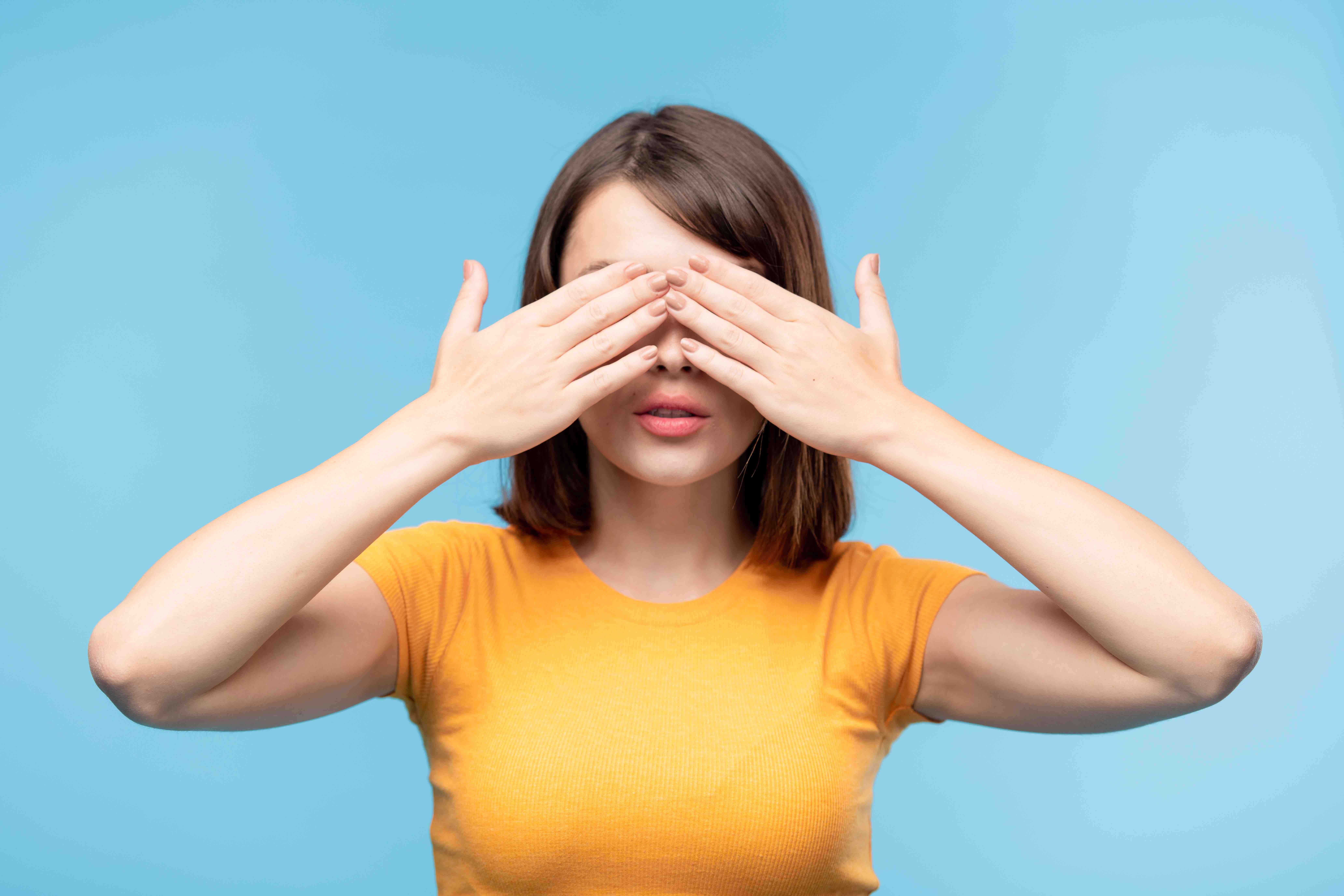 Young brunette female in yellow t-shirt covering her eyes with hands while standing in front of camera in isolation